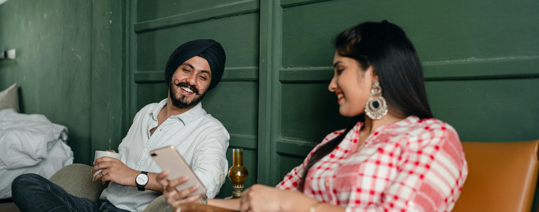 A couple smiling and looking at a phone, enjoying a relaxed moment together in a cozy, green-walled room. The man is holding a cup, and the woman is wearing a red and white checkered shirt with statement earrings.