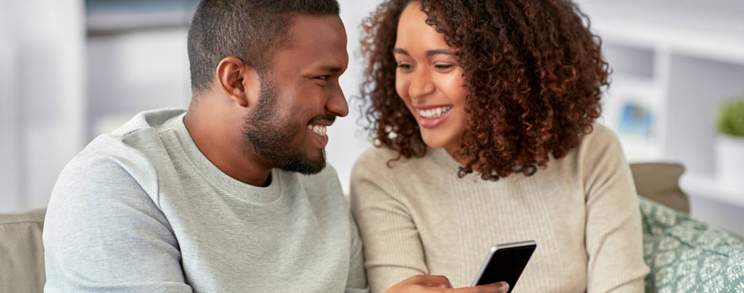 A happy couple smiling and looking at a smartphone together, enjoying each other's company on a cozy couch.