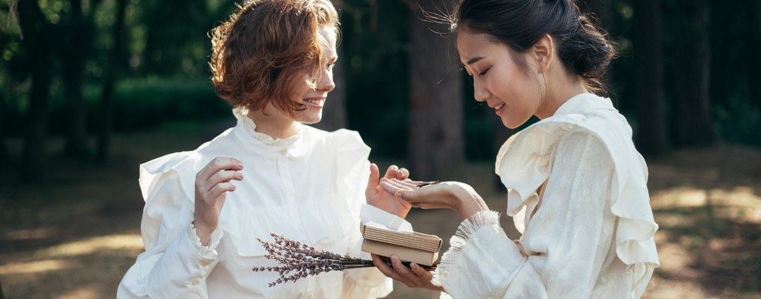 Two women in white dresses, smiling and exchanging a gift outdoors. One woman holds a small box with a lavender sprig, creating a heartfelt and joyful moment.