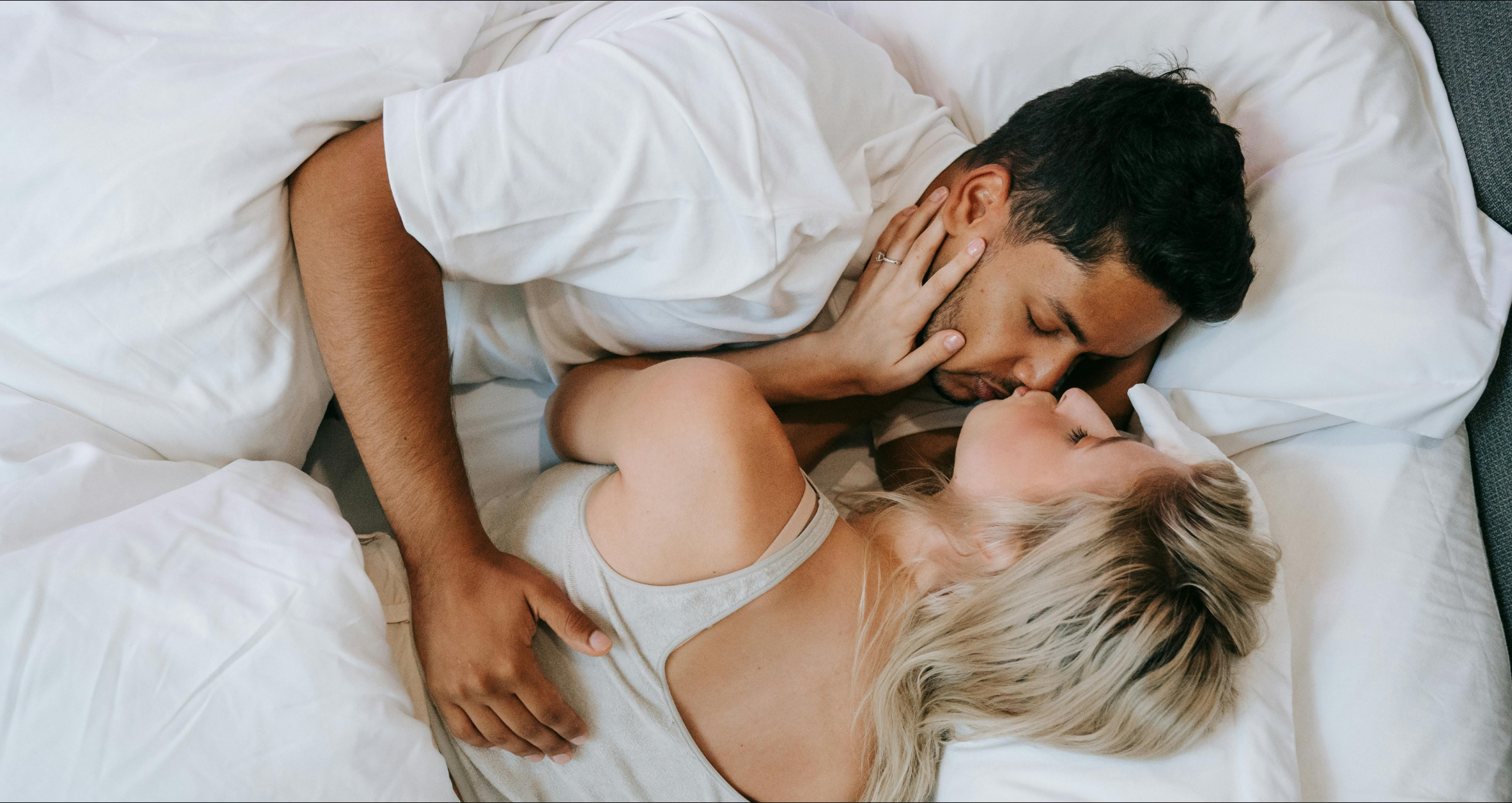 A tender moment between a diverse couple lying in bed, the woman in a grey tank top and the man in a white shirt, holding each other close, with a serene and affectionate expression on their faces, suggesting intimacy and connection in a peaceful bedroom setting