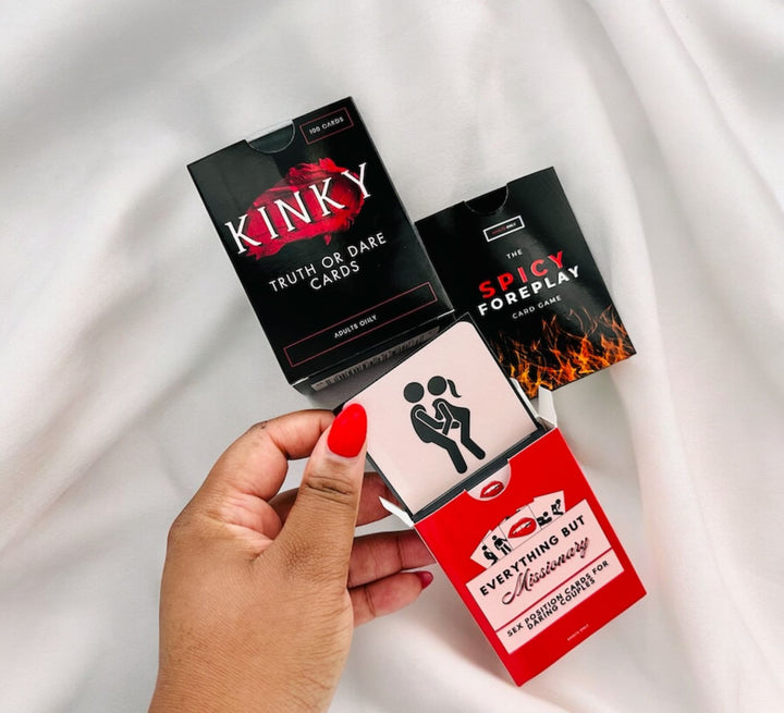 "Adult card games for couples, featuring a hand holding 'Kinky Truth or Dare Cards', 'Spicy Foreplay Card Game', and 'Everything But Missionary' against a white satin background."