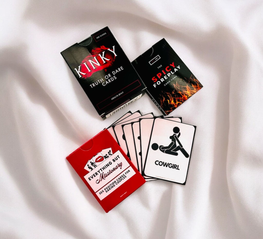 "Adult-themed card games displayed on white satin, including 'Kinky Truth or Dare', 'Spicy Foreplay', and 'Everything But Missionary', with cards showing different positions like 'Cowgirl'."