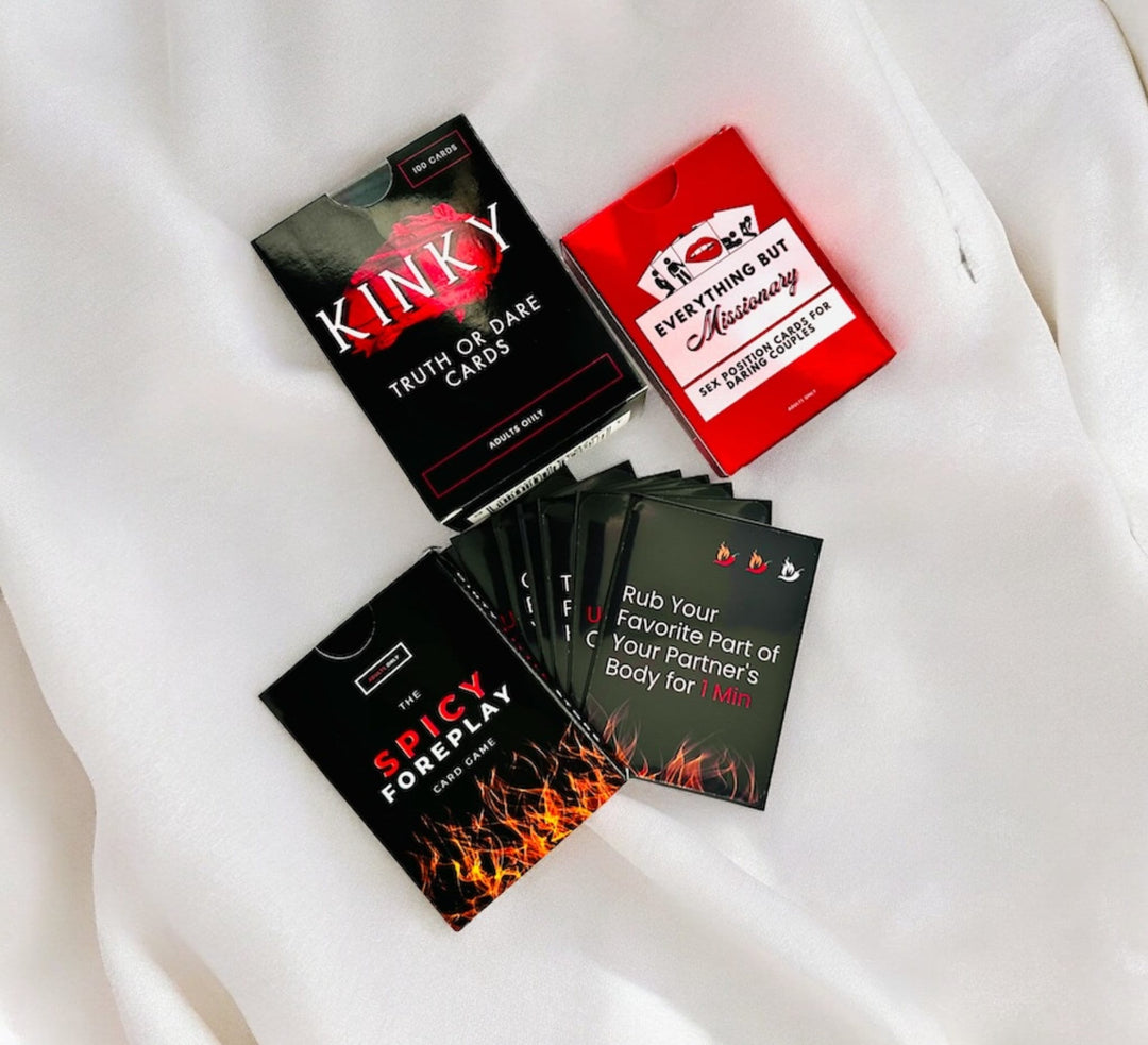 "Adult card games collection on white satin fabric, including 'Kinky Truth or Dare', 'Spicy Foreplay', and 'Everything But Missionary', with a 'Spicy Foreplay' card displaying a playful activity suggestion."