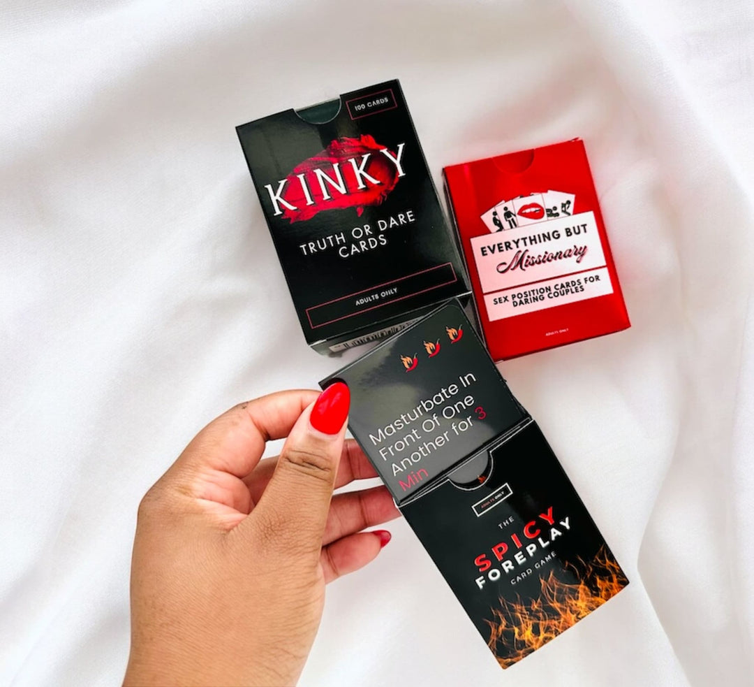 "Hand holding a selection of adult-themed card games on a white satin background, including 'Kinky Truth or Dare', 'Spicy Foreplay', and 'Everything But Missionary', with a visible 'Spicy Foreplay' card suggesting an intimate challenge."