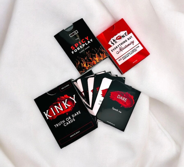 "Variety of adult card games on white satin including 'Kinky Truth or Dare', 'Spicy Foreplay', and 'Everything But Missionary', featuring cards with dares like 'Spank me'."