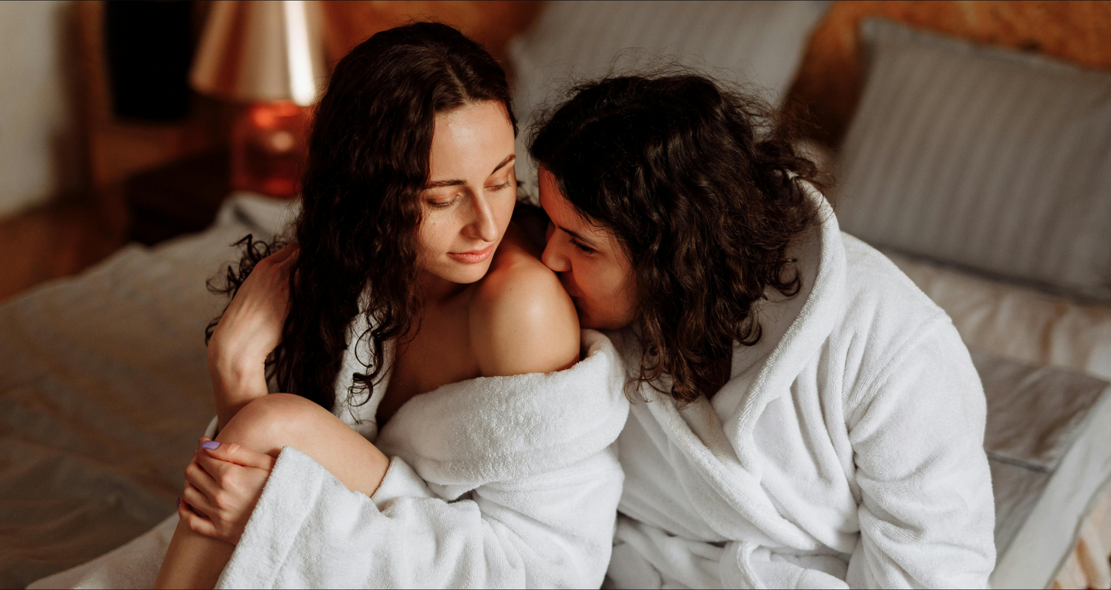 Two women in white bathrobes sharing an intimate moment, embracing and gazing into each other's eyes, conveying a sense of closeness and affection in a cozy, warmly lit bedroom setting.