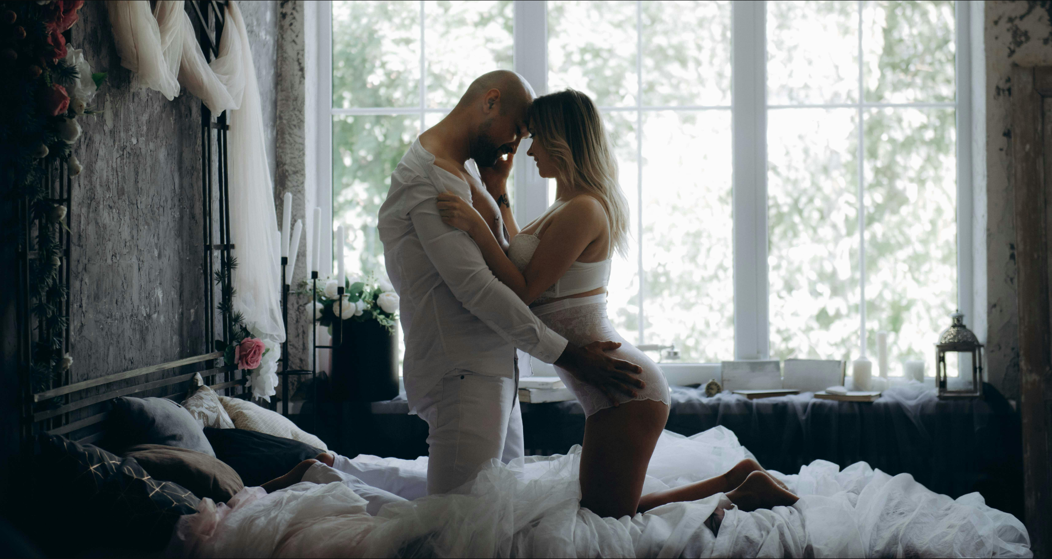 A romantic couple embracing by a window in a room with white bedding and candles.